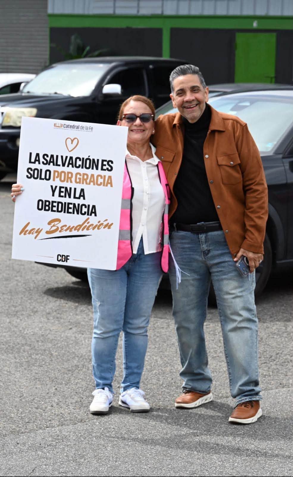 Voluntaria con el Pastor Walter y el letrero La salvación es solo por gracia y en la obediencia hay bendición